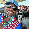man, smiling, sunglasses, cap, dog, black_and_white_dog, car_interior, seat, seatbelt, jacket, stars_and_stripes, flag_pattern, pet, happy, portrait, reflection, window, daylight, face, animal