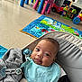baby, infant, cute, playmat, toys, pacifier, seat, blue_bib, gray_clothing, floor, indoor, child, smiling, tongue_out, colorful, playroom, young_child, baby_boy, comfortable, happy