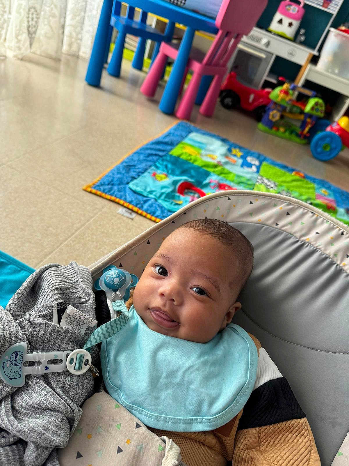 Jésaiah a rejoint le concours — aidez-le/la à gagner de superbes lots ! baby, infant, cute, playmat, toys, pacifier, seat, blue_bib, gray_clothing, floor, indoor, child, smiling, tongue_out, colorful, playroom, young_child, baby_boy, comfortable, happy