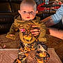 baby, camo_clothing, carpet, child, clothing, cute, eyes, face, indoors, infant, leather_couch, living_room, mouth, person, pink_bow, soft_blanket, standing, supporting_hands, tattooed_hands, warm_lighting