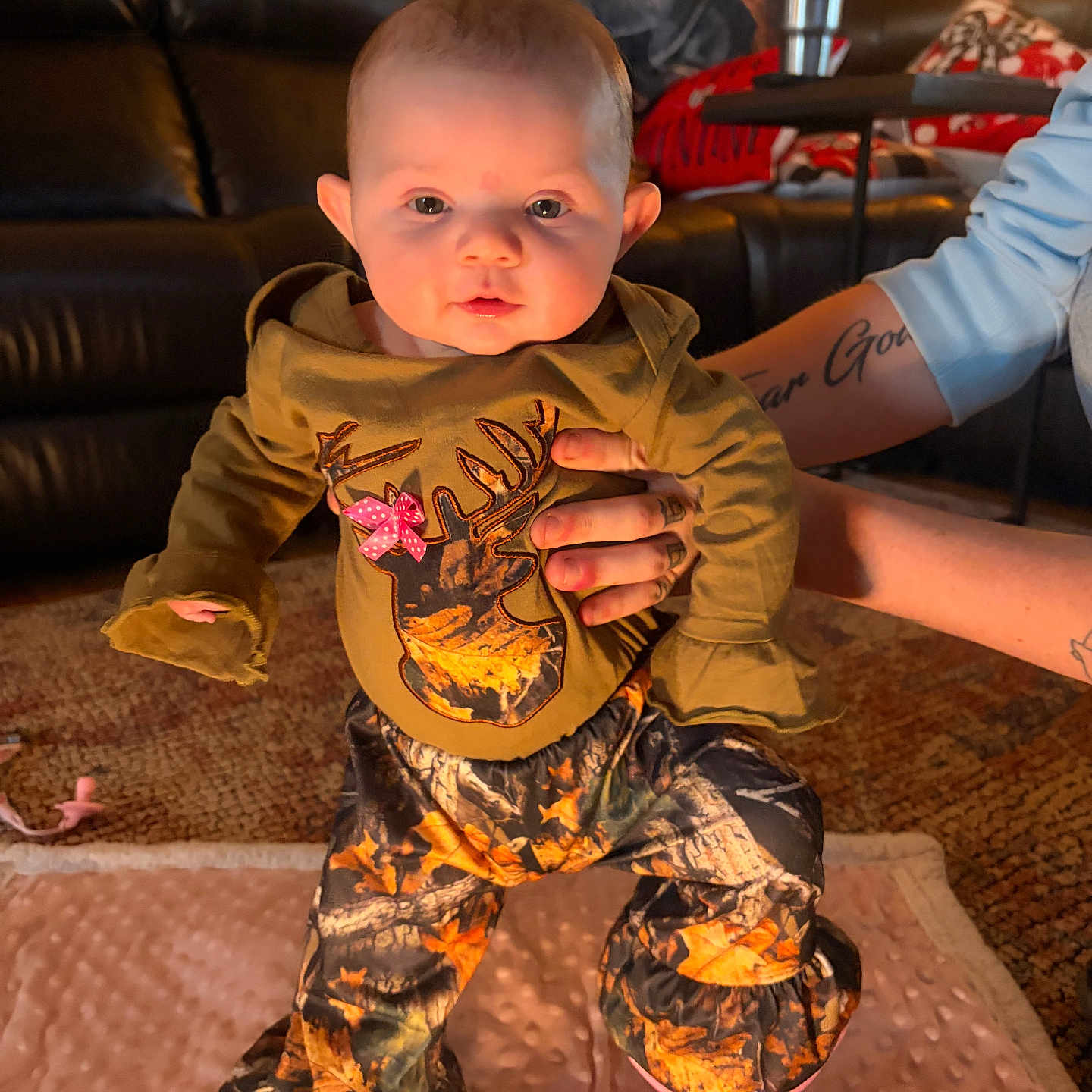 Arya joined the competition — help win amazing prizes! baby, camo_clothing, carpet, child, clothing, cute, eyes, face, indoors, infant, leather_couch, living_room, mouth, person, pink_bow, soft_blanket, standing, supporting_hands, tattooed_hands, warm_lighting