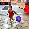 toddler, child, balloon, indoor, red_clothing, purple_balloon, tiles, floor, lockers, counter, red_balloons, bracelet, shoes, person, walk, young_child, hair_bun, toy_store, lighting, smile