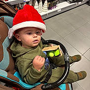 Noan participe au concours pour gagner de l'argent avec cette photo : toddler, child, stroller, red_hat, green_clothing, toy, shopping, store_shelves, holiday_decorations, christmas, ceramic_houses, ceramic_trees, floor_tiles, indoor, person, footwear, hand, curious, shopping_cart, retail