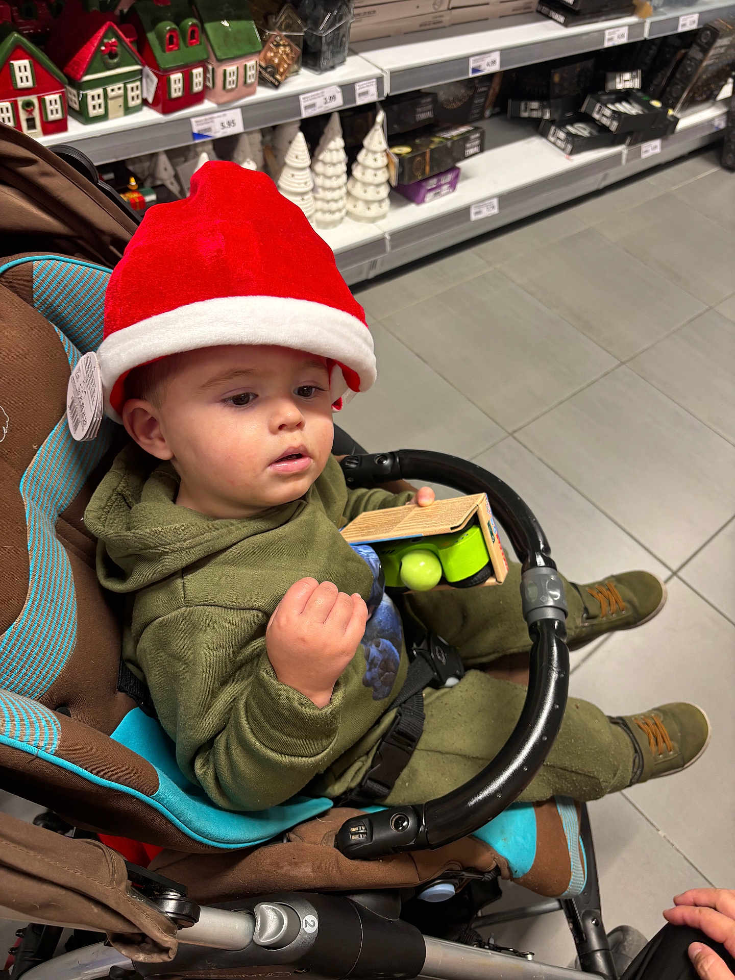 Noan participe au concours pour gagner de l'argent avec cette photo : toddler, child, stroller, red_hat, green_clothing, toy, shopping, store_shelves, holiday_decorations, christmas, ceramic_houses, ceramic_trees, floor_tiles, indoor, person, footwear, hand, curious, shopping_cart, retail