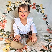 Gianna joined the competition — help win amazing prizes! baby, child, smiling, white_top, ruffled_clothing, hair_bow, gold_shoes, plaid_pants, floral_blanket, sitting, happy, indoors, cute, portrait, infant, person, young_child, bright_eyes, hands, cozy