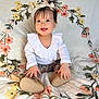 baby, child, smiling, white_top, ruffled_clothing, hair_bow, gold_shoes, plaid_pants, floral_blanket, sitting, happy, indoors, cute, portrait, infant, person, young_child, bright_eyes, hands, cozy