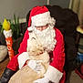 dog, person, santa_claus, red_clothing, white_beard, glasses, indoor, hugging, christmas, holiday, decor, chair, carpet, plant, festive, warmth, affection, gloves, costume, pet