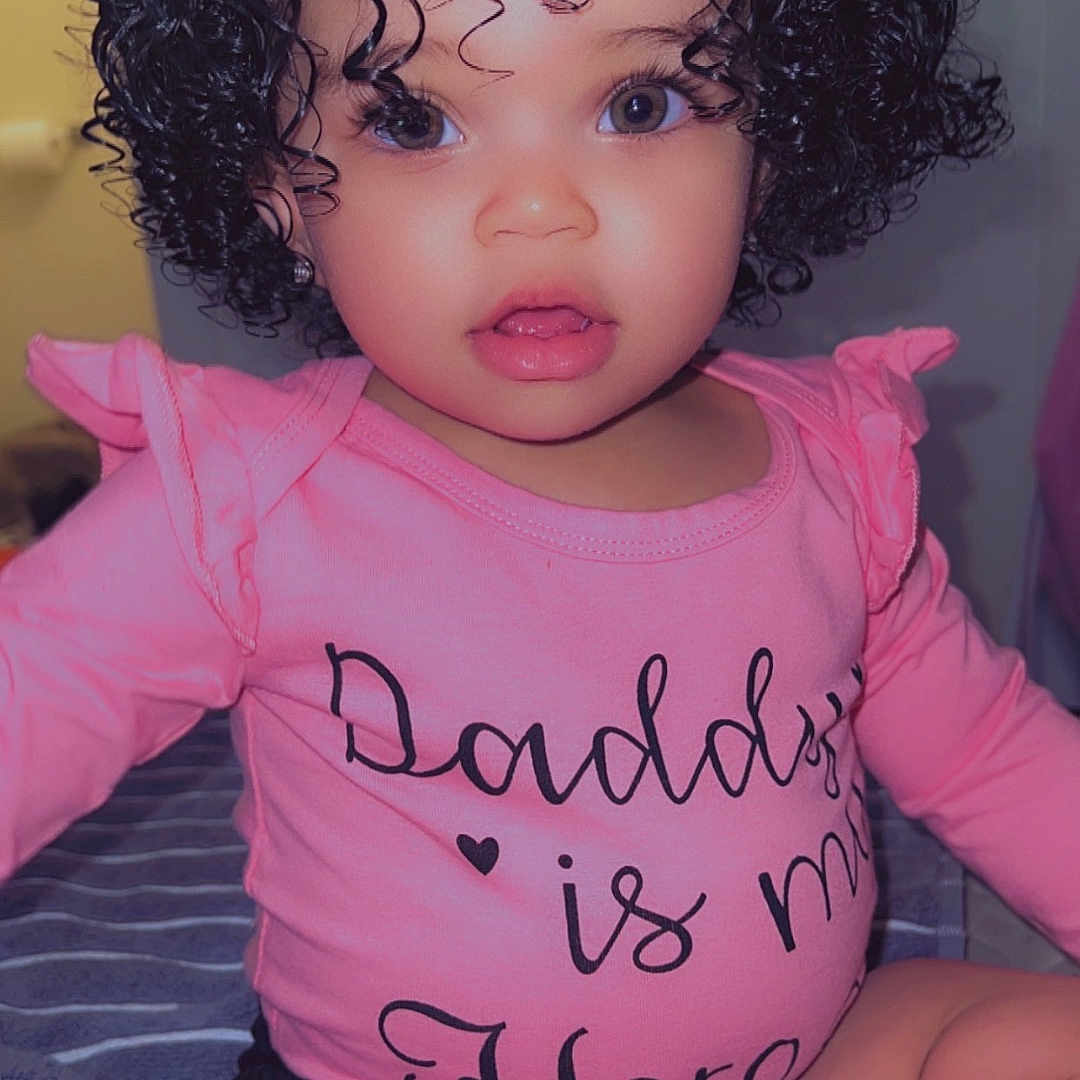 Zariah joined the competition — help win amazing prizes! adorable, baby, blanket, child, clothing, curly_hair, cute, expression, eyes, face, furniture, home, indoor, person, pink_shirt, portrait, sitting, smile, toddler, young_child