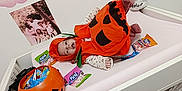 Kiarah a rejoint le concours — aidez-le/la à gagner de superbes lots ! baby, candy, celebration, changing_table, clothing, costume, cute, decorations, face, halloween, holiday, indoor, infant, lying_down, orange, person, plastic_bucket, pumpkin_costume, skull_mask, toy