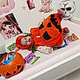 baby, pumpkin_costume, halloween, candy, changing_table, skull_mask, orange, costume, infant, holiday, cute, indoor, toy, plastic_bucket, decorations, face, person, lying_down, clothing, celebration