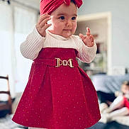 Leyna participe au concours pour gagner de l'argent avec cette photo : baby, child, red_dress, headband, bow, indoor, living_room, white_tights, cute, smiling, standing, soft_lighting, furniture, blurred_background, person, toddler, warm_colors, cozy, portrait, happy