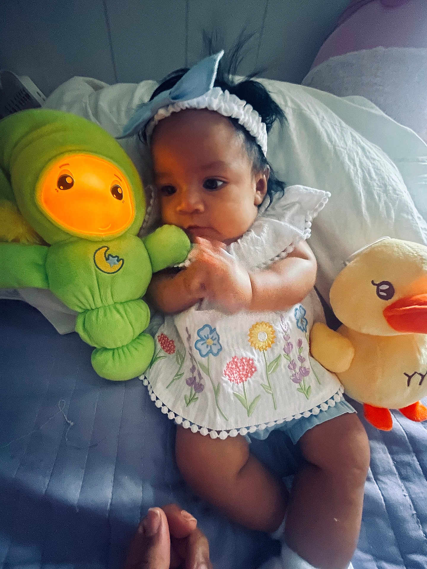 Amelia is registered to the contest to win money with this photo: baby, child, plush_toy, green_toy, yellow_duck, bed, white_headband, floral_clothing, hand, indoor, cute, infant, soft_toy, colorful, lying_down, comfort, person, portrait, expression, cozy