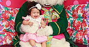 Nala joined the competition — help win amazing prizes! baby, child, easter_bunny, mascot, costume, pink_dress, headband, flower, smiling, happy, floral_background, colorful, purple_petals, green_coat, ribbon, 2026, indoor, portrait, seated, celebration