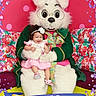 baby, child, easter_bunny, mascot, costume, pink_dress, headband, flower, smiling, happy, floral_background, colorful, purple_petals, green_coat, ribbon, 2026, indoor, portrait, seated, celebration