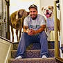 bond, carpet, casual_clothing, dog, friendly, handrail, happy, hat, home, indoor, man, nuzzling, pet, railing, relaxed, sitting, smiling, stairs, tattoo, tongue_out