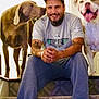 animal, brown_dog, cap, carpet, casual_clothing, companionship, dog, facial_hair, friendship, happy, home, indoor, man, pets, sitting, smiling, stairs, tattoos, tongue_out, white_dog