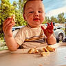 baby, car, child, clothing, daylight, expression, face, food, hands, highchair, messy, outdoor, patio, person, sandwich, sauce, shirt, toddler, tray, trees