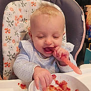 Sophia is registered to the contest to win money with this photo: baby, blond_hair, bowl, child, clothing, cute, eating, food, happy, high_chair, indoor, infant, kitchen, magnet, messy, person, playful, refrigerator, smiling, table