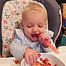 baby, high_chair, food, bowl, kitchen, smiling, happy, blond_hair, child, indoor, eating, magnet, refrigerator, clothing, table, messy, cute, infant, playful, person