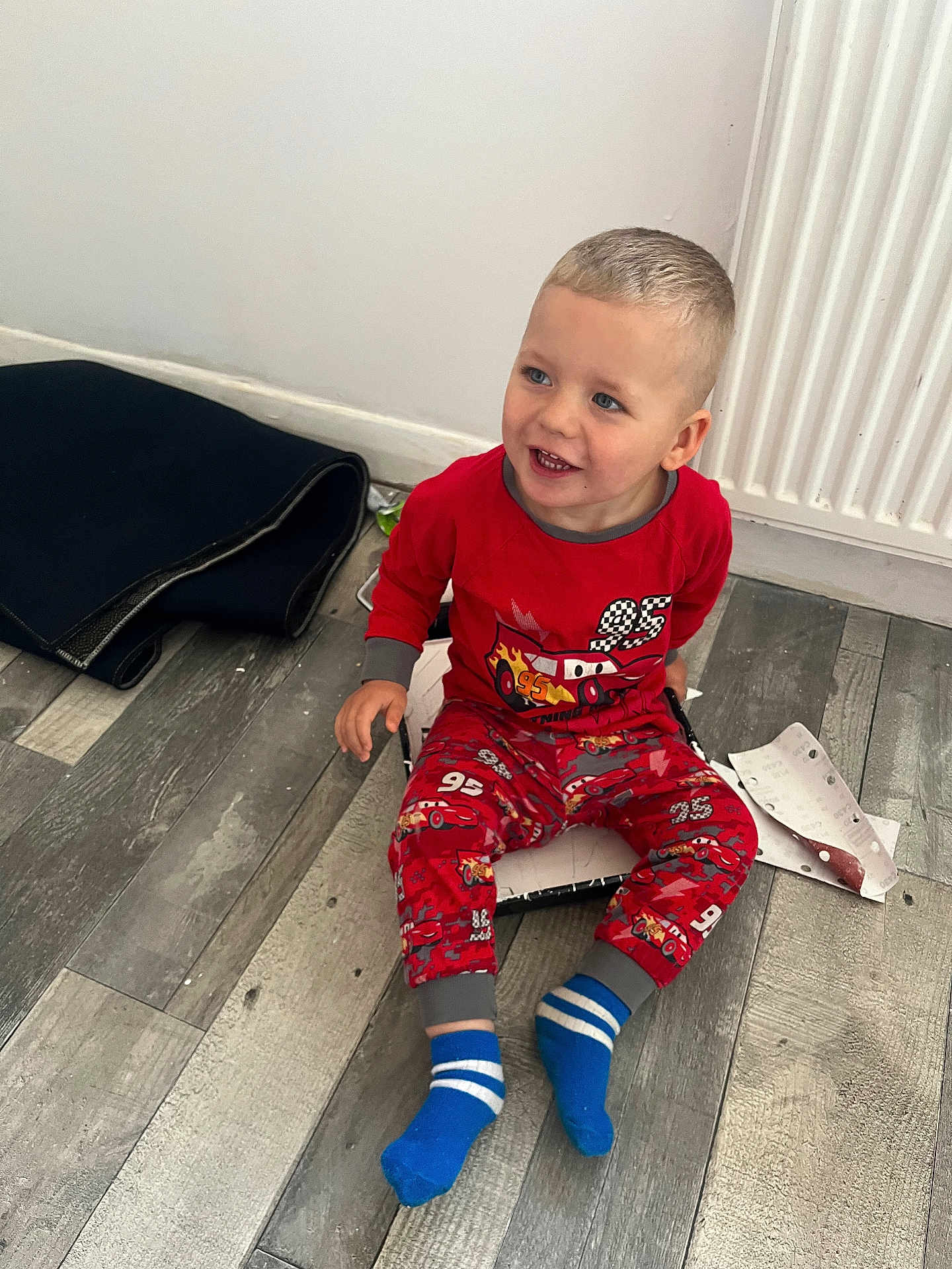 Matas Gluskovas joined the competition — help win amazing prizes! toddler, child, pajamas, red_clothing, sitting, floor, wooden_floor, smiling, blue_socks, indoors, radiator, paper, mat, playful, happy, cute, person, young_child, casual, home