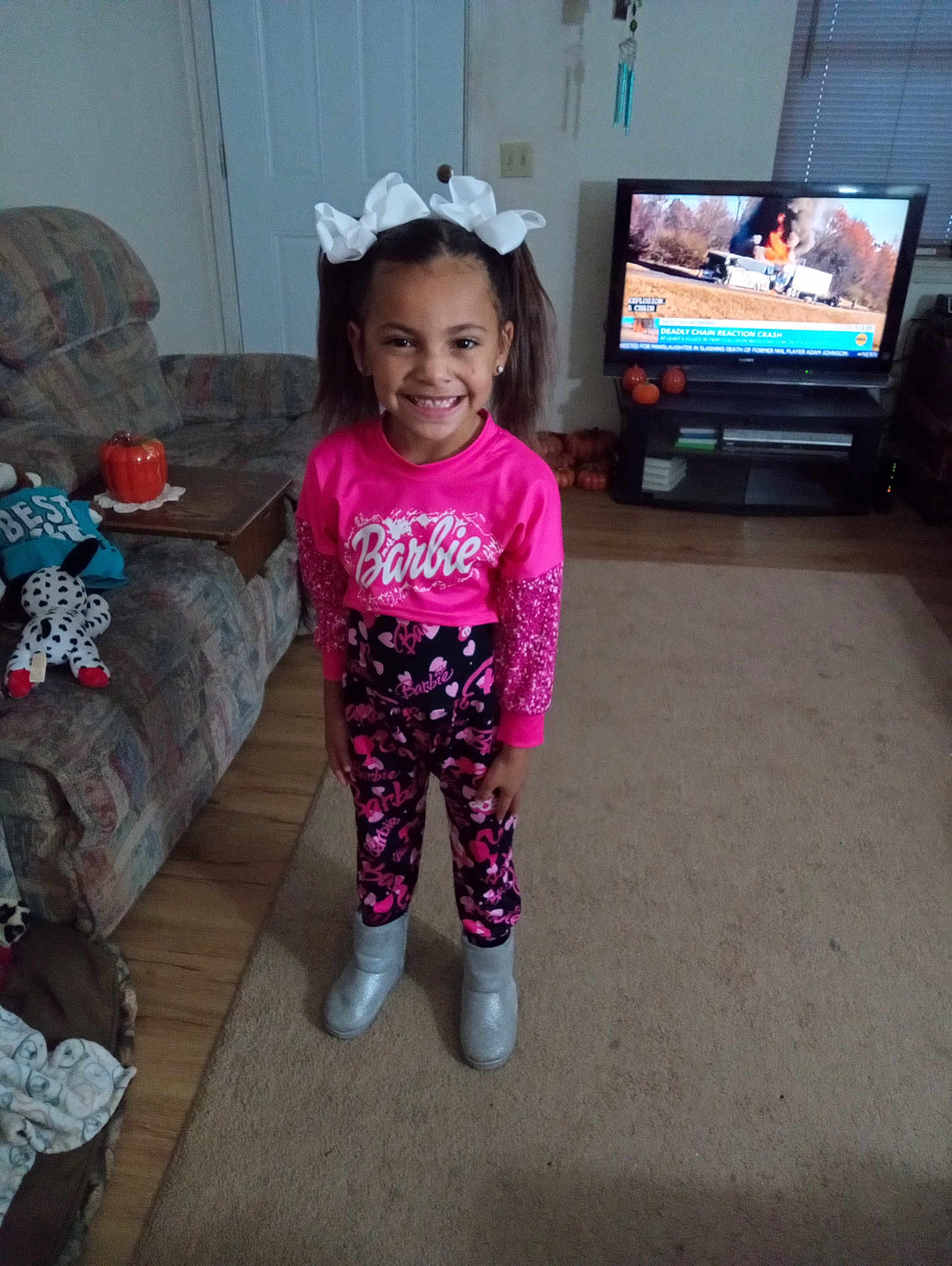 Ma'Kiah is registered to the contest to win money with this photo: child, girl, smiling, standing, indoors, living_room, television, pink_clothing, boots, pumpkins, couch, carpet, door, toy, hair_bows, news_broadcast, wooden_floor, furniture, decor, happy