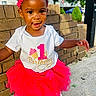 child, toddler, birthday, pink_tutu, headband, happy, outdoor, brick_wall, sidewalk, white_shoes, smiling, cute, celebration, person, young_child, sunlight, fashion, playful, festive, one_year_old