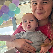Carson is registered to the contest to win money with this photo: baby, child, woman, balloons, tongue_out, indoor, window, smile, holding, casual_clothing, person, portrait, happy, light, hair, hand, table, celebration, cute, fun