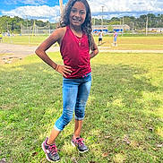 Celeste is registered to the contest to win money with this photo: child, girl, smiling, standing, grass, outdoor, daytime, casual_clothing, jeans, tank_top, sneakers, sports_field, sky, clouds, trees, people_in_background, electric_pole, fence, sidewalk, sunlight