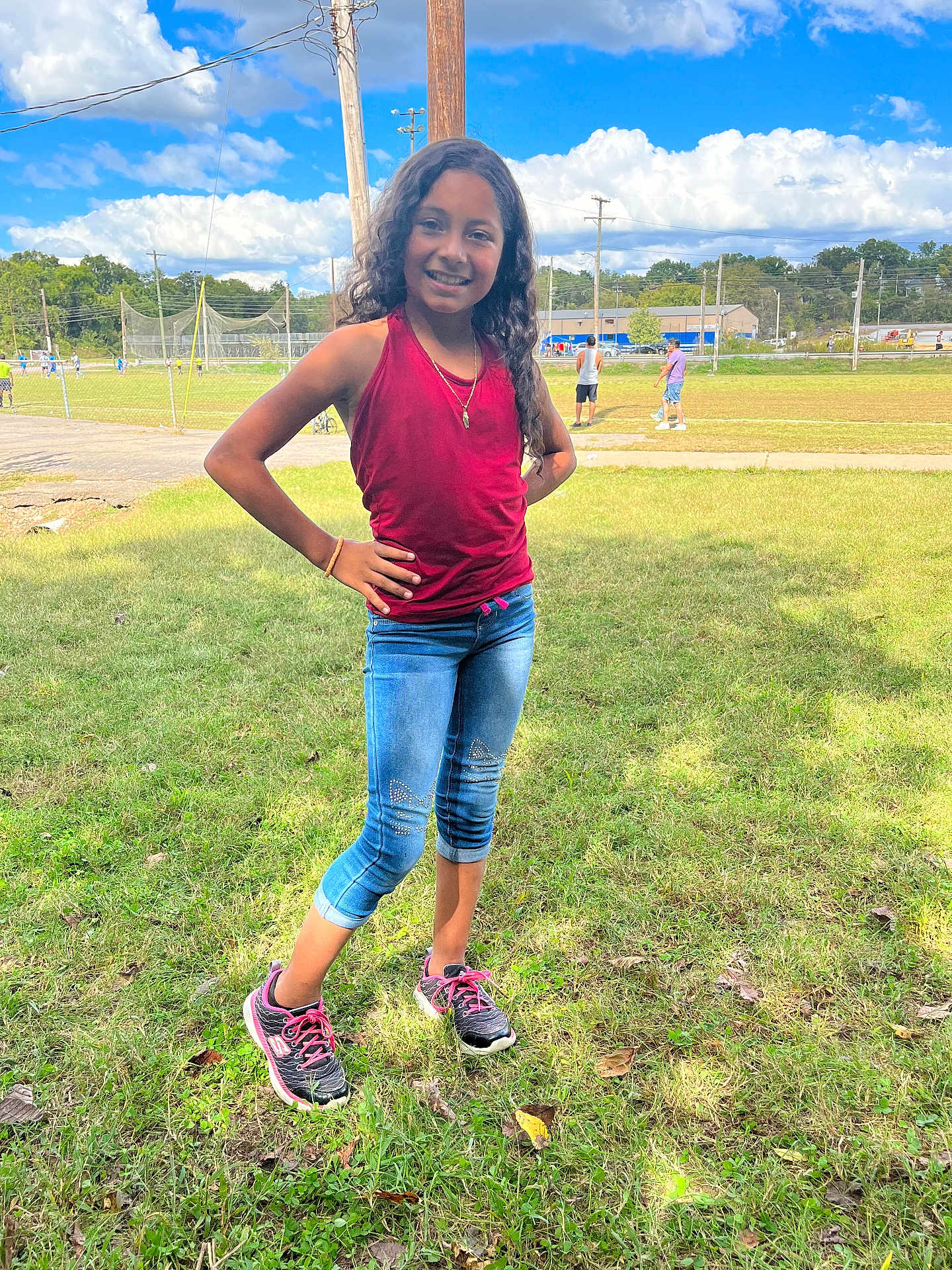 Celeste is registered to the contest to win money with this photo: child, girl, smiling, standing, grass, outdoor, daytime, casual_clothing, jeans, tank_top, sneakers, sports_field, sky, clouds, trees, people_in_background, electric_pole, fence, sidewalk, sunlight