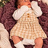 baby, blanket, bouquet, bunny, child, cozy, crocheted_toy, dress, face, flowers, gingham, indoors, infant, lying_down, newborn, nursery, portrait, smiling, socks, soft_lighting
