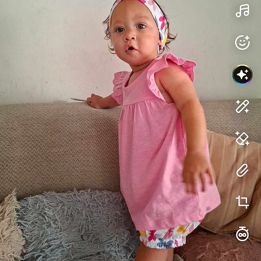 Nayaah participe au concours pour gagner de l'argent avec cette photo : app_interface, button, casual, child, colorful_shorts, couch, curly_hair, expression, furniture, headband, indoor, music_icon, person, phone_screenshot, pink_dress, portrait, rug, standing, toddler, wall