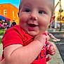 baby, infant, smile, closeup, portrait, outdoor, blue_sky, red_shirt, blue_shorts, hand, parent_hand, building, street_fair, vendor_tent, lamp_post, cheeks, eyes, happy, candid, person