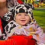 baby, infant, child, smiling, cow_hat, cow_print, blanket, red_glove, parent, partial_face, outdoor, night, park, grass, bokeh_lights, cheerful, portrait, jacket, face, cute