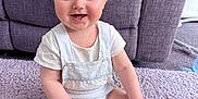 Finley is registered to the contest to win money with this photo: baby, child, smiling, sitting, blonde_hair, blue_eyes, white_clothing, socks, carpet, rug, couch, pillows, indoor, cute, happy, young_child, infant, portrait, home, soft_texture
