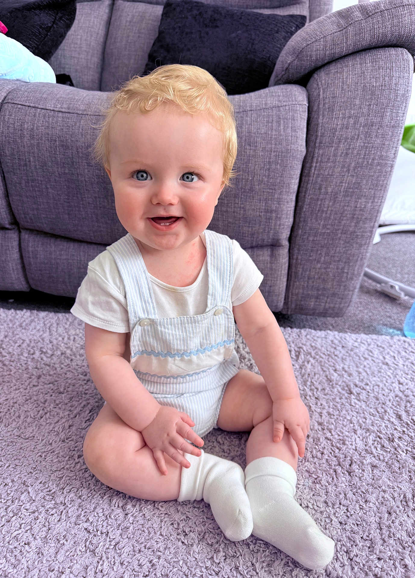 Finley is registered to the contest to win money with this photo: baby, child, smiling, sitting, blonde_hair, blue_eyes, white_clothing, socks, carpet, rug, couch, pillows, indoor, cute, happy, young_child, infant, portrait, home, soft_texture