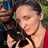 black_cat, cat, closeup, dreadlocks, face, glasses, happy, hat, holding, man, nature, outdoor, pet, portrait, selfie, smile, sunlight, trees, woman, yellow_eyes
