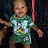baby, child, infant, smiling, portrait, sitting, green_outfit, mickey_mouse, cartoon, shirt, shorts, black_background, plush_blanket, earrings, happy, indoor, photograph, adult_hand, fashion, closeup
