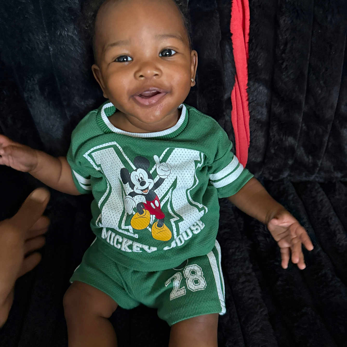 Kylan is registered to the contest to win money with this photo: baby, child, infant, smiling, portrait, sitting, green_outfit, mickey_mouse, cartoon, shirt, shorts, black_background, plush_blanket, earrings, happy, indoor, photograph, adult_hand, fashion, closeup