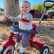 Jacob is registered to the contest to win money with this photo: toddler, child, tricycle, outdoor, sunny, happy, smiling, tree, vehicle, grass, play, toy, clothing, vest, pants, socks, handlebars, wheels, daytime, nature