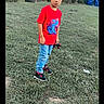 child, boy, grass, park, toy_car, red_tshirt, jeans, sneakers, trees, outdoor, nature, greenery, recycling_bins, casual_clothing, young, standing, playing, daylight, curious, portrait