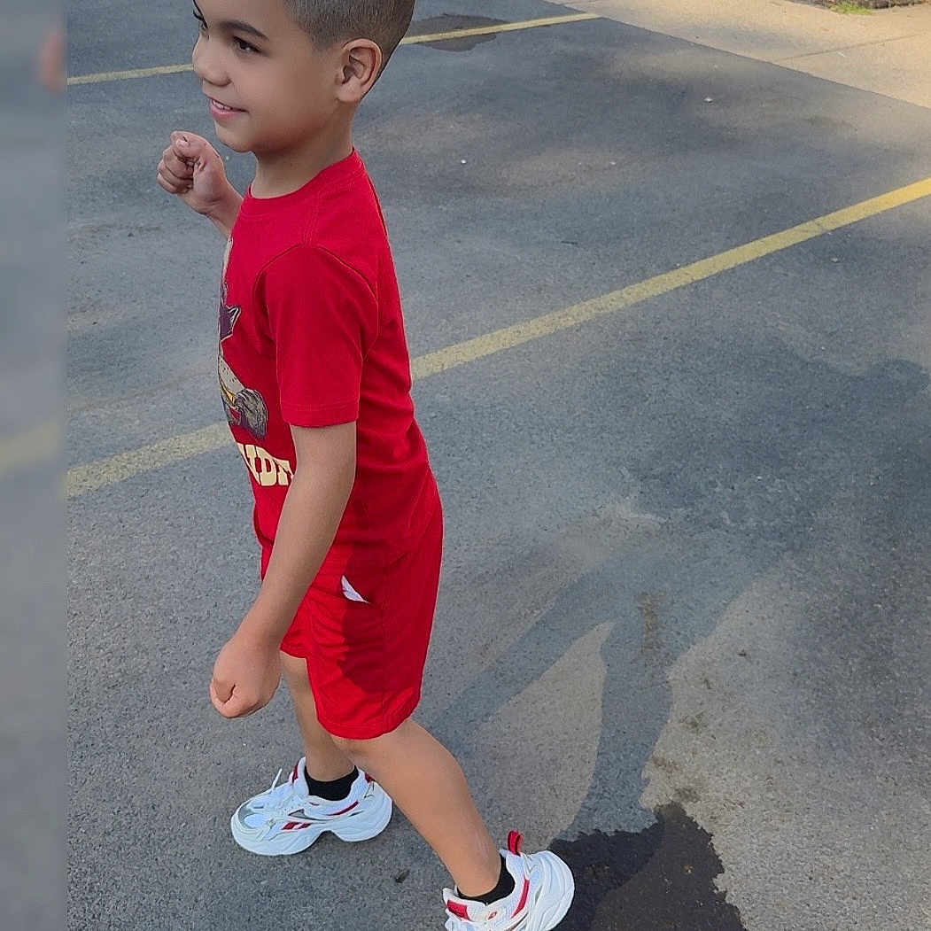 Xadiel Estévez joined the competition — help win amazing prizes! child, boy, red_clothing, shorts, sneakers, white_shoes, outdoor, parking_lot, asphalt, shadow, smiling, short_hair, curly_hair, side_view, casual, daytime, playful, active, young, person