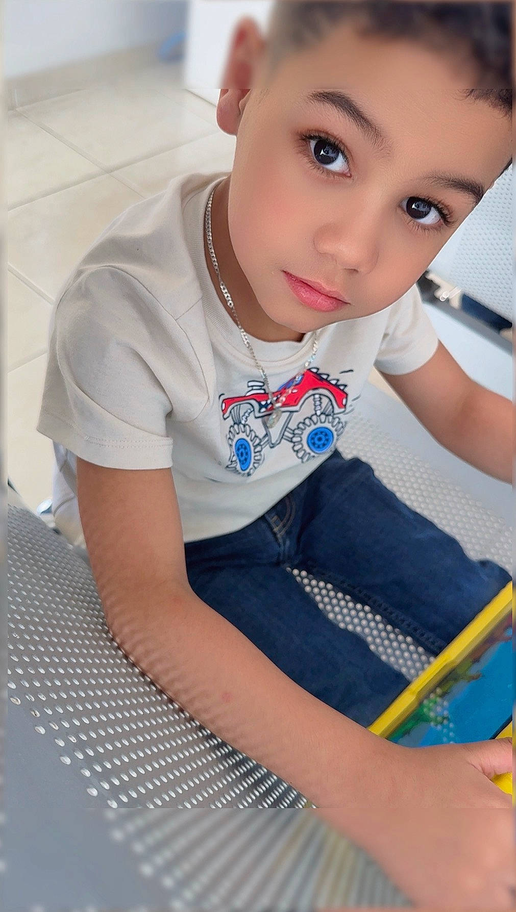 Xadiel Estévez is registered to the contest to win money with this photo: bench, boy, casual_clothing, child, eyes, face, floor, indoor, jeans, looking_up, metal_bench, necklace, portrait, short_hair, sitting, tablet, tile_floor, toy, white_tshirt, young