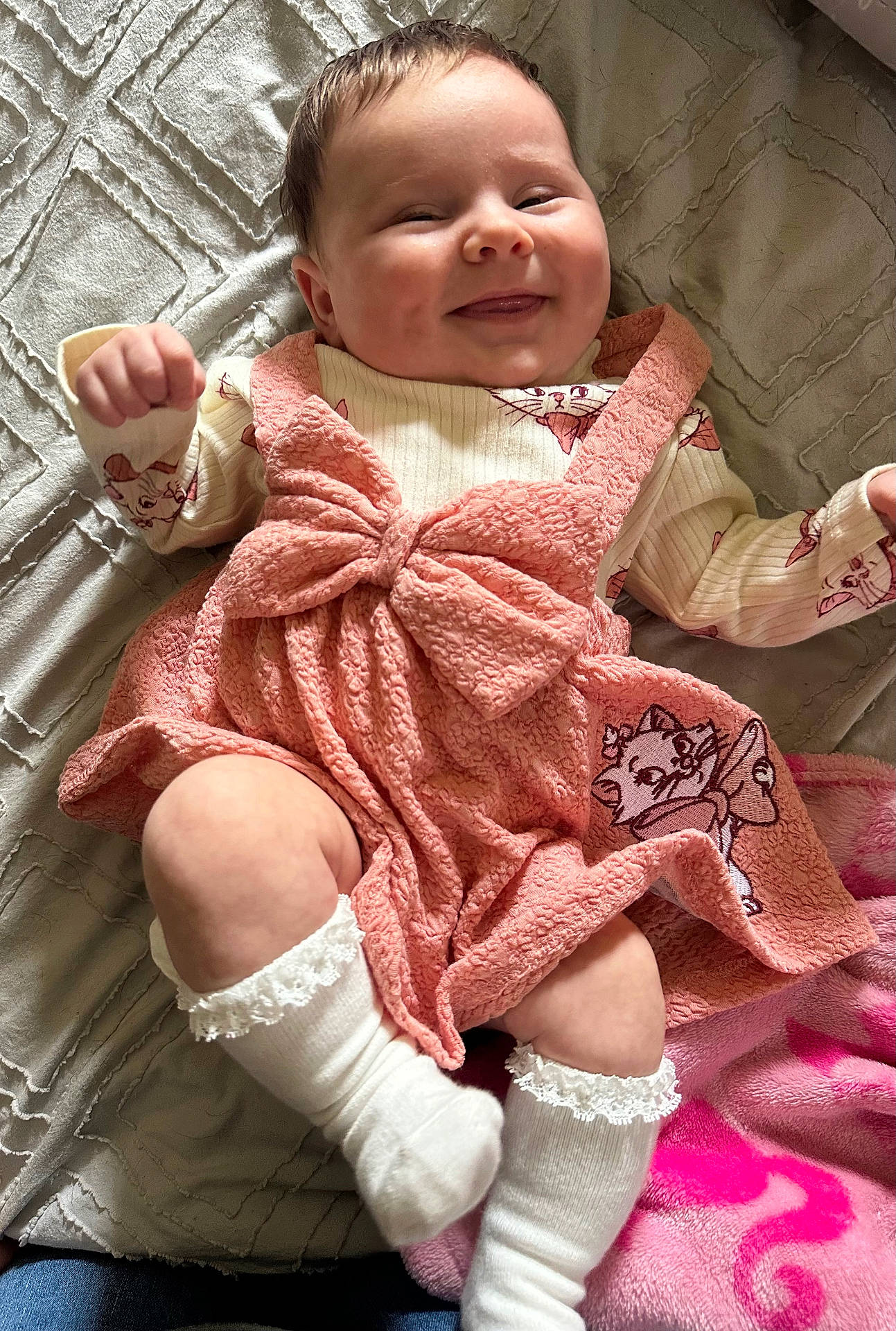 baby, blanket, bow, child, clothing, cute, face, hand, happy, indoor, infant, legs, lying_down, patterned, person, pink_dress, quilt, smile, soft_texture, white_socks