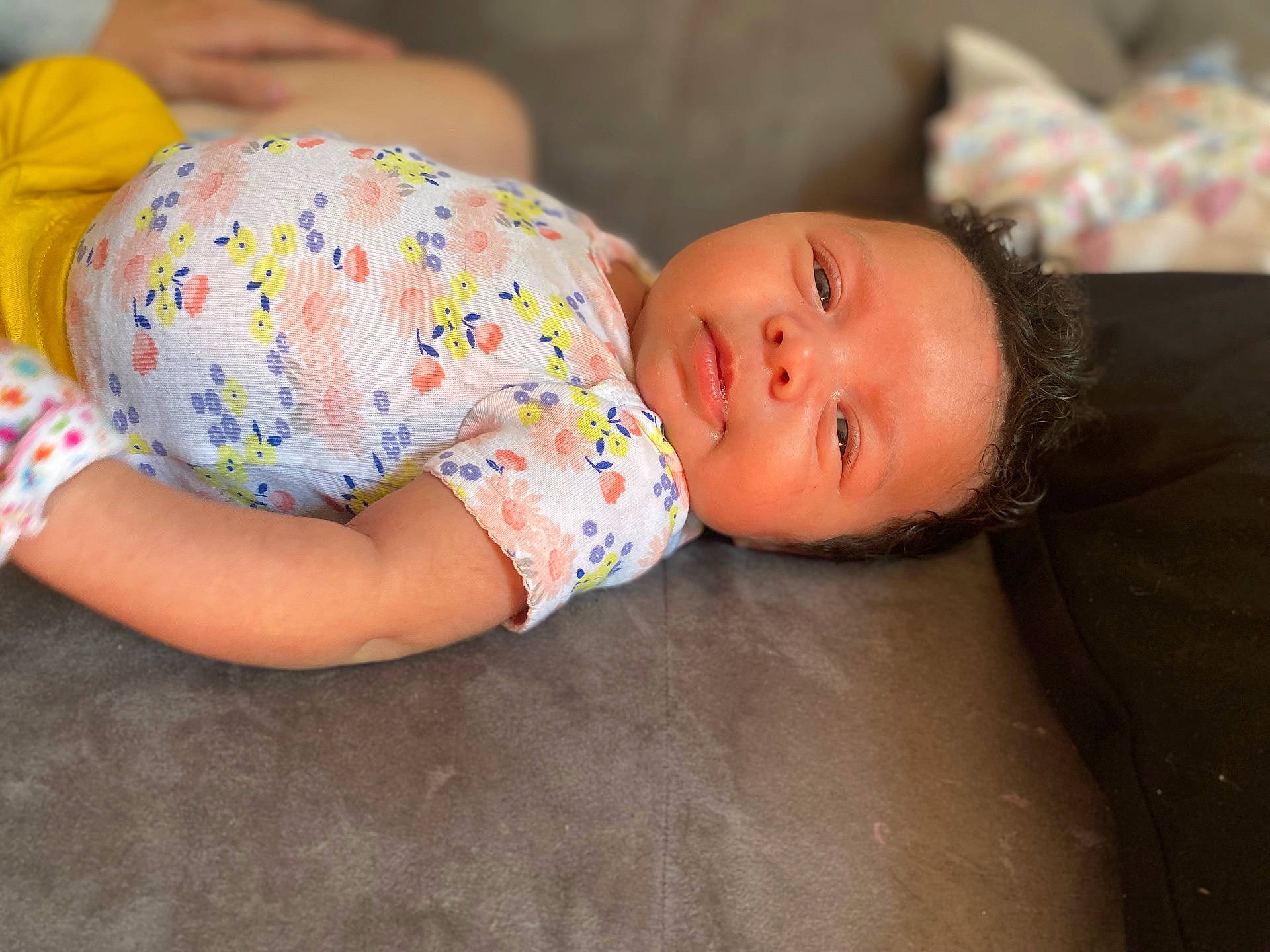 arm, baby, casual, child, couch, curly_hair, cute, expression, face, floral_clothing, human, indoors, infant, lying_down, portrait, resting, skin, soft_surface, yellow_pants, young