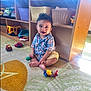 basket, carpet, child, clothing, colorful, cute, floor, furniture, hair, indoors, natural_light, person, play, playroom, plush_toy, sitting, smiling, toddler, toy, wooden_shelf