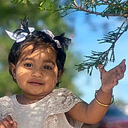 Zunairah is registered to the contest to win money with this photo: child, toddler, girl, pigtails, bows, white_dress, lace, bracelet, outdoor, tree_branch, leaf, sunlight, shadow, blue_sky, portrait, smile, cute, face, hand, daylight