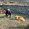 animal, bone, casual_clothing, chewing, crouching, daylight, dog, fluffy_dog, grass, hill, man, mountain, nature, old_man, outdoor, pet, rock, rural, sky, stone_wall