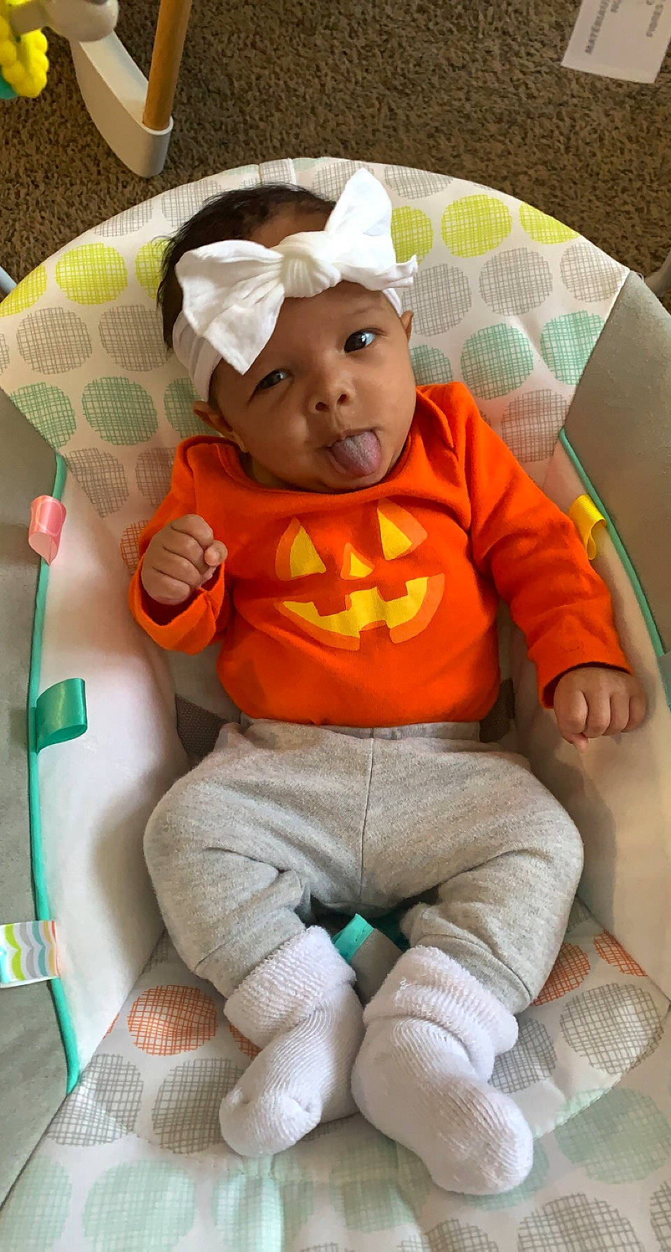 Avah is registered to the contest to win money with this photo: baby, baby_products, baby_toddler_clothing, chair, cheek, child, comfort, face, head, headwear, lap, linens, orange, person, personal_protective_equipment, pumpkin, sitting, sleeve, sock, t_shirt