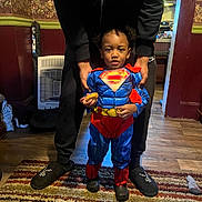 Kashton joined the competition — help win amazing prizes! child, superman_costume, cookie, adult_hands, rug, carpet, indoor, wood_floor, wallpaper, heater, slippers, socks, kitchen, doorway, person, standing, costume, curly_hair, home, casual