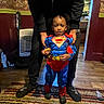 child, superman_costume, cookie, adult_hands, rug, carpet, indoor, wood_floor, wallpaper, heater, slippers, socks, kitchen, doorway, person, standing, costume, curly_hair, home, casual